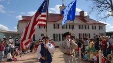Two paraders holding flags high above their heads and dressed in colonial garb including hats walk down a paved trail in front of George Washington's Mount Vernon in Virginia, USA. Around them are spectators in modern-looking clothes at this reenactment.