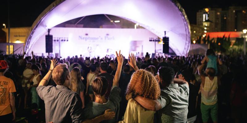 A group of people among a crowd stand at an outdoor concert where the stage is lit up with lavender lights in Virginia Beach, Virginia, USA.