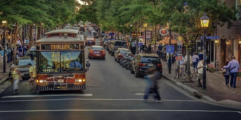 A bustling, tree-covered King Street in Old Town Alexandria, USA near dusk. People walk along the cobblestone streets in front of shops under golden-lit streetlamps. The King Street Trolley is stopped on the road, welcoming people in and out of the bus.