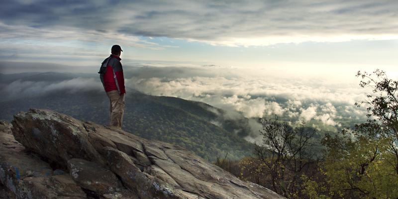 A hiker stands on Humpback Rock overlooking an overcast, but beautiful sky, streaked with wispy clouds and peaks in the distance