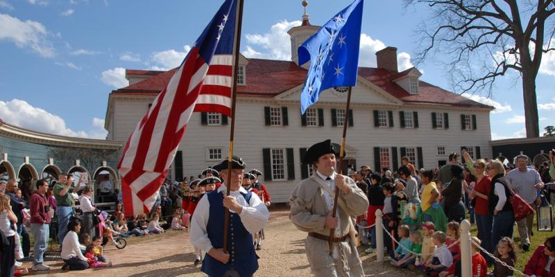 Two paraders holding flags high above their heads and dressed in colonial garb including hats walk down a paved trail in front of George Washington's Mount Vernon in Virginia, USA. Around them are spectators in modern-looking clothes at this reenactment.