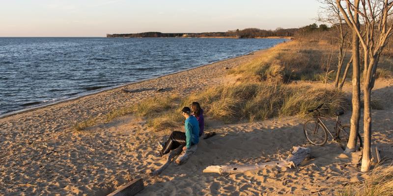 A couple sits on the beach next to wind-blown sea grass as they look out at the water in Kent Island< Maryland, USA