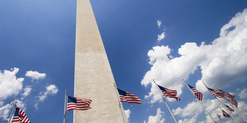 A picture of the Washington Monument from the ground looking up towards the blue, cloud-dotted sky. There are American flags flying around the base of the monument