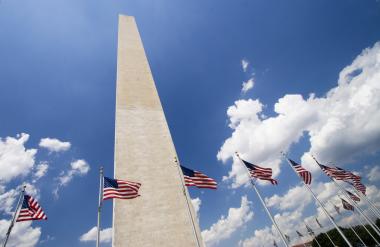 A picture of the Washington Monument from the ground looking up towards the blue, cloud-dotted sky. There are American flags flying around the base of the monument