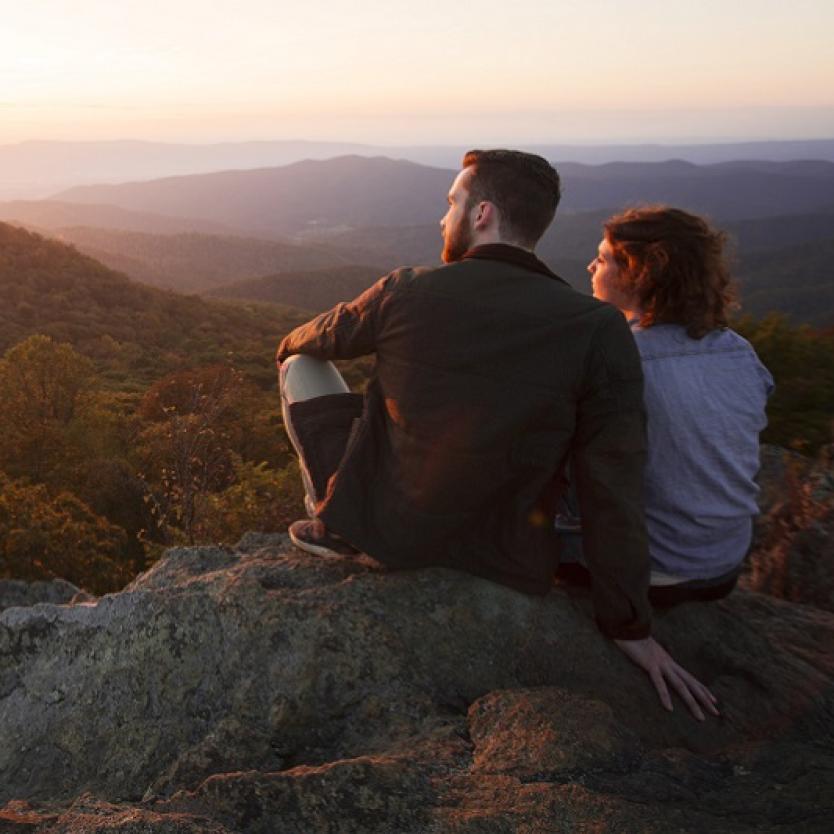 Two serene-looking people sit side-by-side on large rocks overlooking the hills and forests of Shenandoah National Park in Virginia, USA at sunset. The glow of the sun reflects on their faces.
