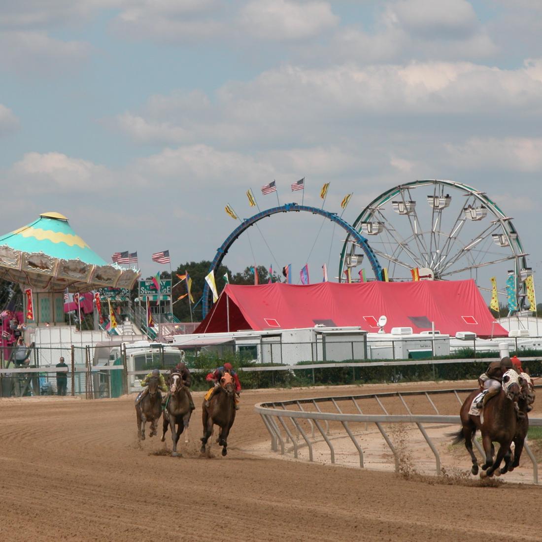 Horses race on a track with fair rides in the background